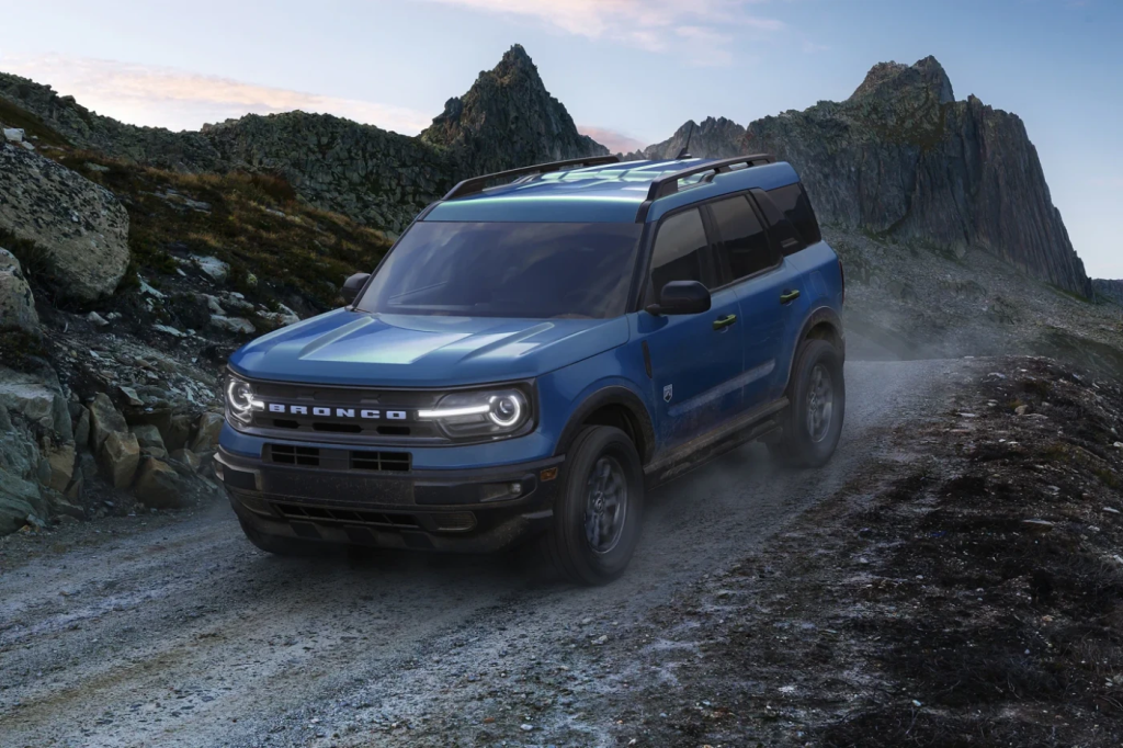 A blue SUV drives on a rugged mountain road under a clear sky. Dust trails behind, highlighting its off-road capability and adventurous spirit.
