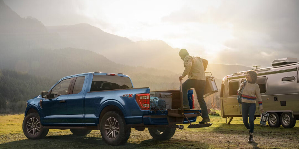 A blue pickup truck parked on a grassy field with mountains in the background at sunrise. One person steps off the truck bed with a cooler, while another walks holding a lantern. An Airstream trailer is nearby. The mood is serene and adventurous.