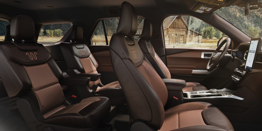 Luxurious SUV interior with brown leather seats, spacious layout, and a modern dashboard featuring a large touchscreen. Rustic cabin visible outside.