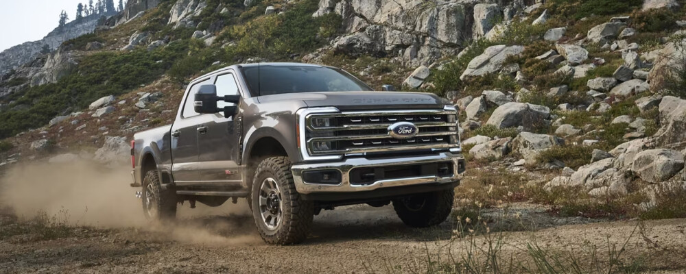 A rugged pickup truck kicks up dust as it drives off-road on a rocky mountain path, surrounded by lush greenery and rugged cliffs under a clear sky.