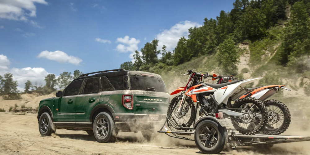 A green SUV drives on a dusty road pulling a trailer with two dirt bikes. Pine trees and a clear sky in the background convey adventure and freedom.