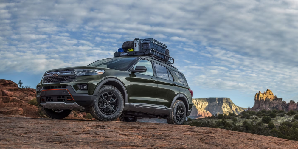 Green SUV packed with gear on a red rock ledge, set against a dramatic, cloudy sky and distant desert cliffs, evoking adventure and exploration.