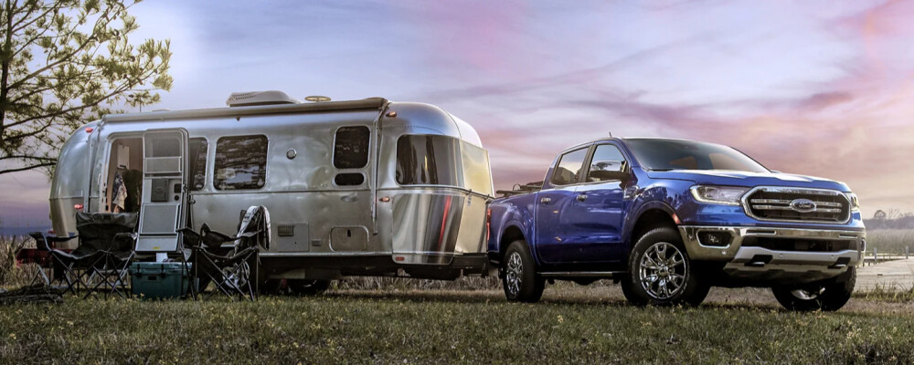 A blue pickup truck is parked next to a silver Airstream trailer in a grassy field at sunset. Camping chairs and trees are visible, conveying a peaceful outdoor setting.