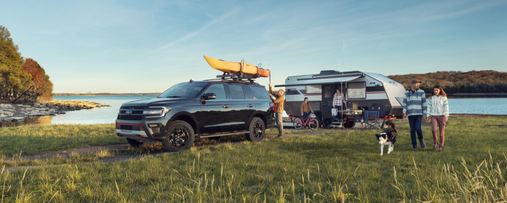 A couple and a dog walk near an SUV and camper by a lakeside. Two kayaks are on the SUV roof, and a family prepares by the camper in a serene, sunny setting.