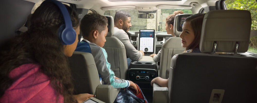 A family of five sits in an SUV, looking relaxed and happy. The kids in the backseat engage with tablets and headphones, while the parents converse up front.