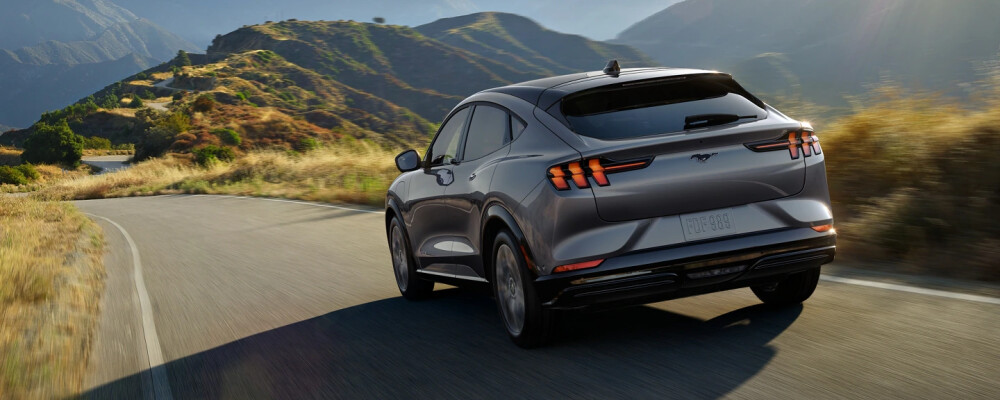 A sleek silver SUV drives on a winding mountain road under a clear sky. Sunlight highlights the rolling hills and adds a sense of freedom and adventure.