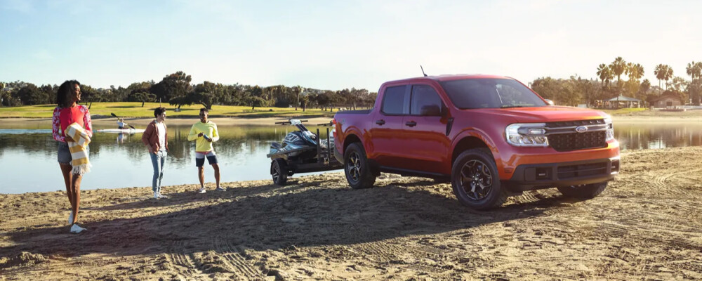 A red pickup truck towing a jet ski is parked on a sandy lakeshore. Three people in casual attire chat nearby. Bright, sunny day; relaxed atmosphere.
