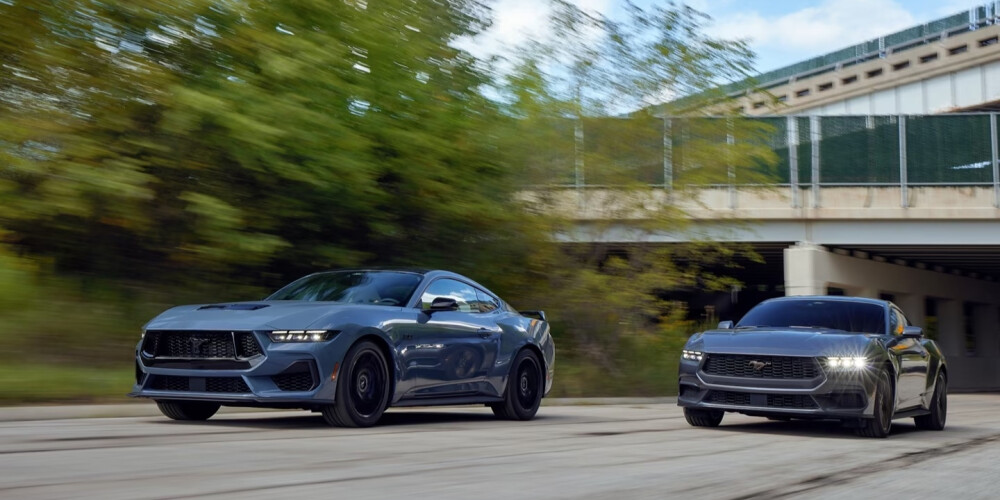 Two sleek, modern muscle cars drive side by side on a road near a bridge, with blurred green trees in the background, conveying speed and power.