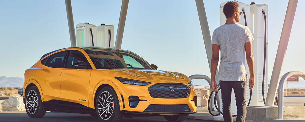 A person stands at an electric vehicle charging station beside a bright yellow car. The scene is sunny and open, conveying a modern and eco-friendly vibe.