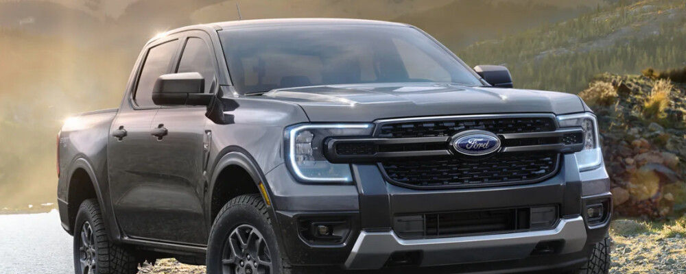 Silver pickup truck with bold grille and rugged tires is parked by a reflective lake, mountain backdrop. The scene conveys strength and adventure.