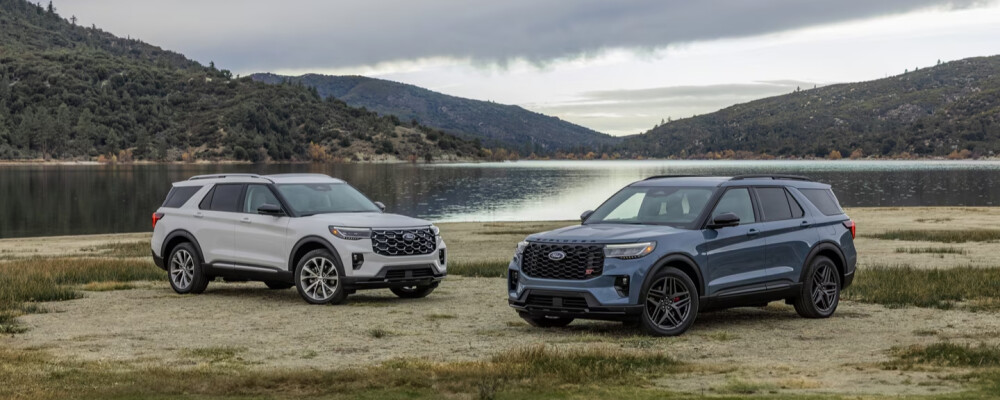Two SUVs parked on a grassy shoreline by a tranquil lake, surrounded by rolling hills and a cloudy sky, convey a peaceful and adventurous mood.