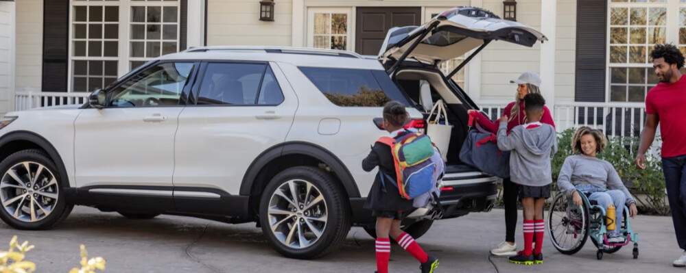 A family packs a white SUV outside a house. Two kids with backpacks and a woman load bags, while a person in a wheelchair smiles nearby. A man watches.