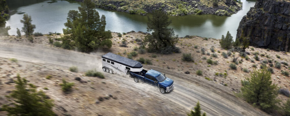 A blue truck pulls a white trailer on a dusty road beside a scenic lake surrounded by trees and rocky hills, evoking a sense of adventure and freedom.